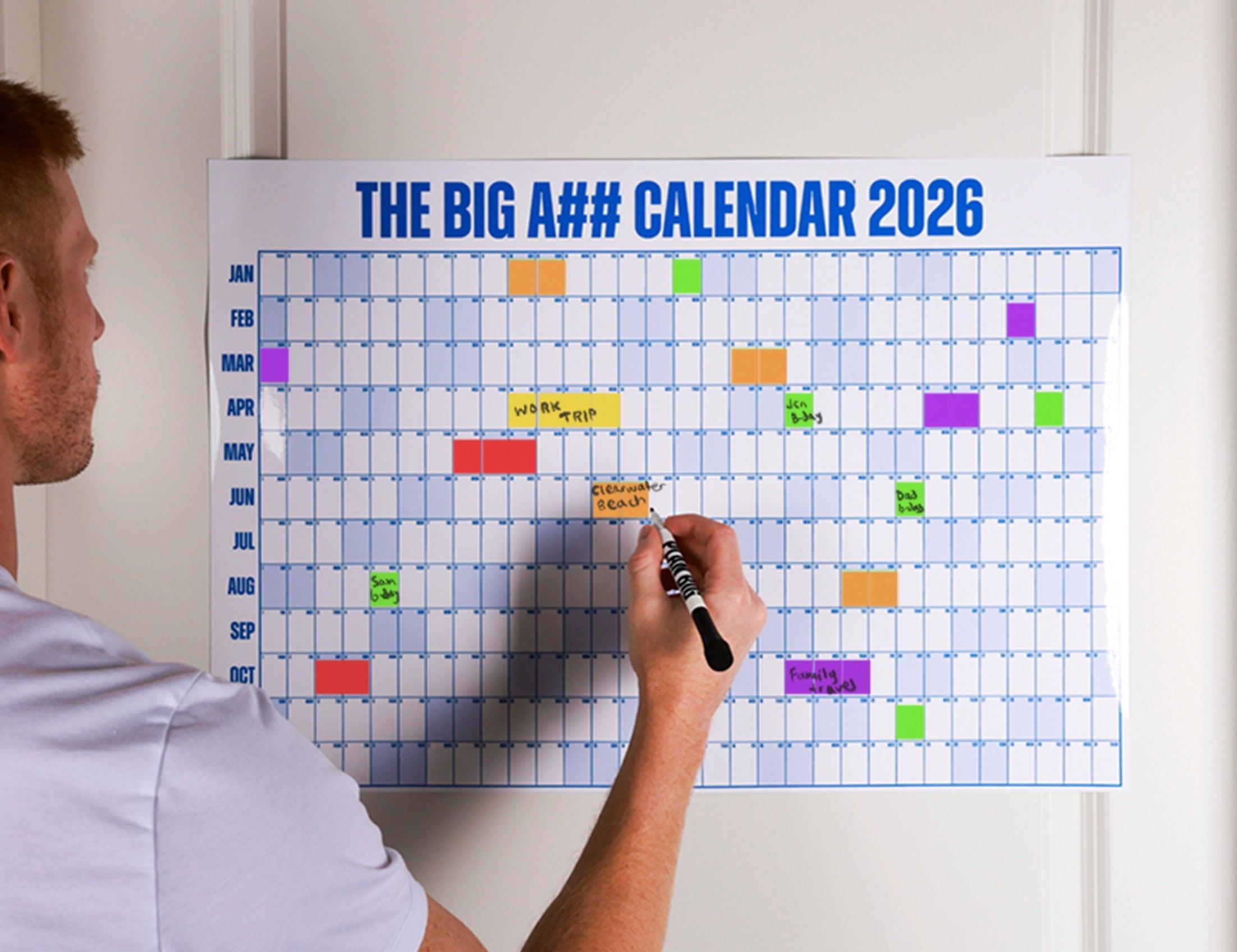 Person writing on a colorful calendar labeled 'THE BIG A#$# CALENDAR 2026' with a marker.